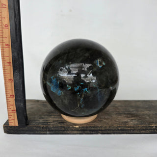 Labradorite crystal ball with blue and white interior on a wooden stand with ruler for scale against a white background